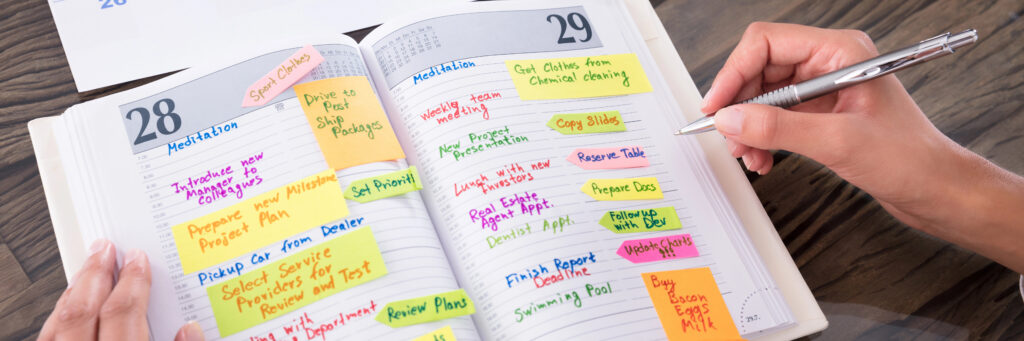 Photograph of an open planner with multiple appointments and notes per day, a hand with a pencil hovers above it as if they are about to add more.
