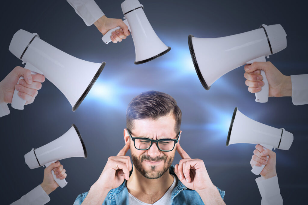 a man in the lower center of the image has his fingers in his ears against the 5 megaphones aimed at him from the sides and the top.