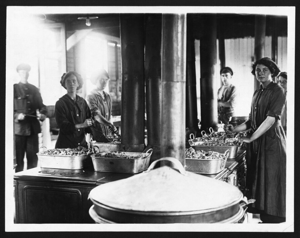 Black & White photograph of 5 women cooks at the front lines during WWI, they are preparing large roasting pans heaped with foods A soldier stands in the background.