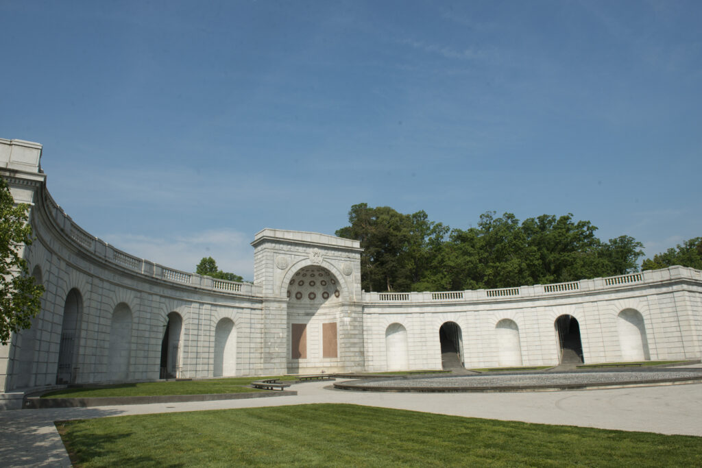 Color photograph of the Military Women's Memorial a building of white stone in an open arch facing the drive, in the center of the building is a tall arch with memorial placques inside it. On each side of that are 5 smaller arched doorways.