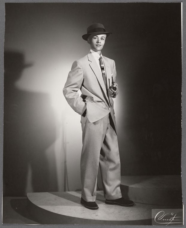 Black and white publicity shot of Storme standing in her male persona (no mustache). Her hair isn't entirely visible but gives the impression of being light in color. she is wearing a dark fedora, has on a boxy style light colored two piece suite. She holds a pipe in her left hand and has her right hand in her pocket.