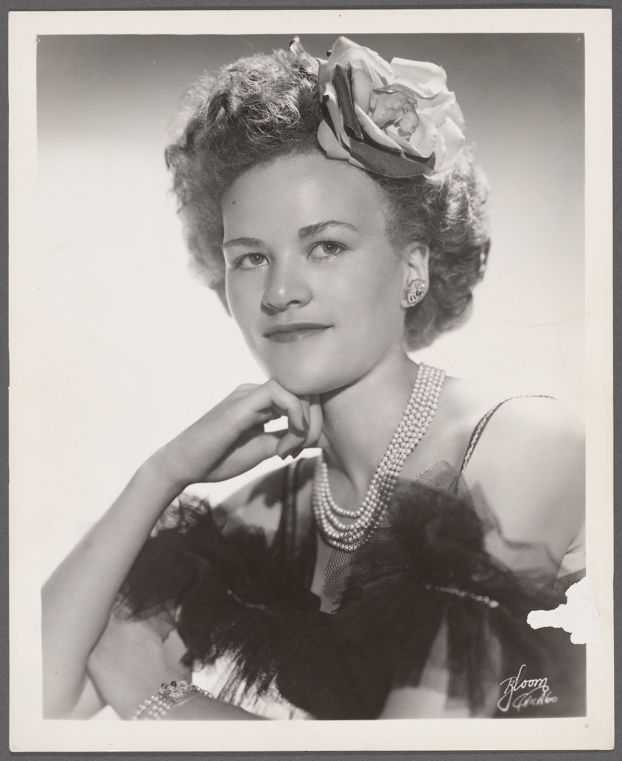 Professional headshot photograph of Stormé she is wearing a cloth flower in her hair, earrings and a multi-strand pearl necklace with a spagetti-strap dress with black tulle ruffles across the neckline just above the bust