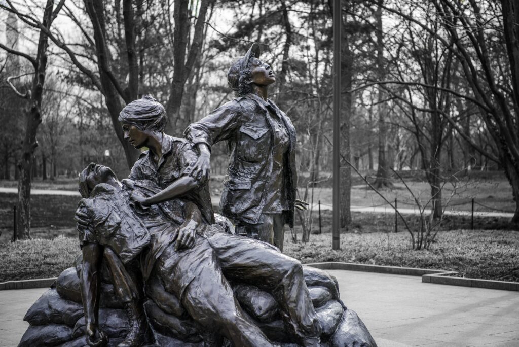 Black and white photograph of the Vietnam Women's Memorial in Washington DC shows two nurses and an injured soldier. One nurse holds the soldier who is limp in her arms. Her hand pressed over his heart. The second woman stands behind them, looking toward the sky, her hand grips the upper arm of the first woman.