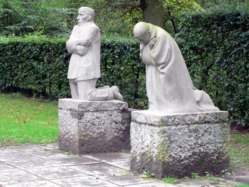 The image is a color photo of the sculpture, The Grieving Parents. Consisting of two statues on rough cut stone blocks depicts Karl Kollwitz on his knees, face frozen in grief and arms hugging himself tightly on one block. Käthe is on her knees, her head bowed, her arms crossed over her in a position of deep grief.