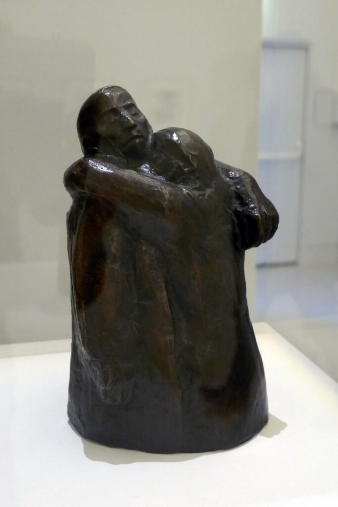 photograph of a small sculpture in a museum. The sculpture shows a man and woman from the head to their hips, The woman's head is nestled against the neck of a man whose head is back and his eyes are closed. she has her arms wrapped around the man's neck, her face nestled between his neck and shoulder. Their bodies almost blend together.