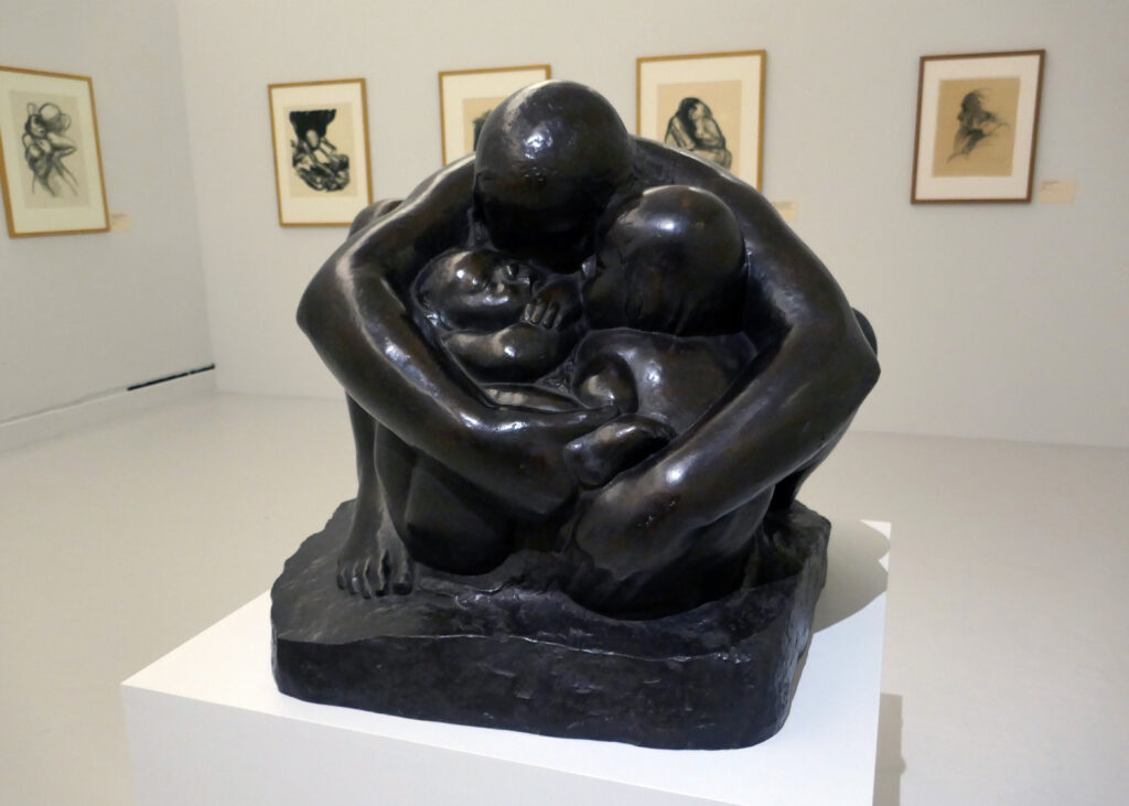 Photograph of a bronze statue in a museum. The statue is of a woman squatting, feet flat on the ground, head over the top of, and arms wrapped around an infant and small child who are also protected by her legs.