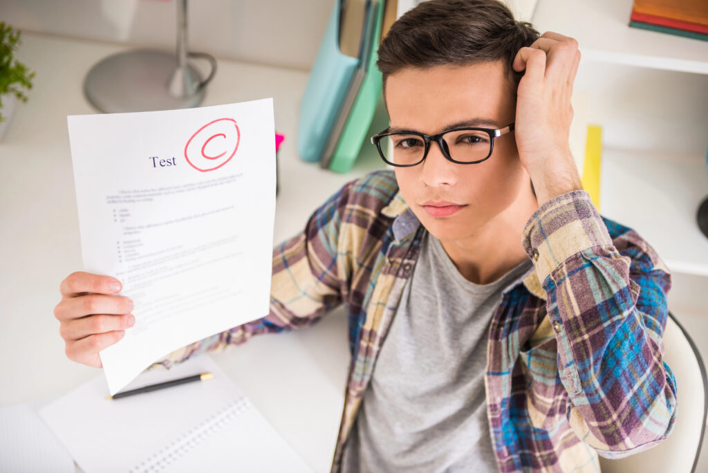 Color Photo of a school aged boy scratching his head in unhappy puzzlement while holding up a test with a red C at the top