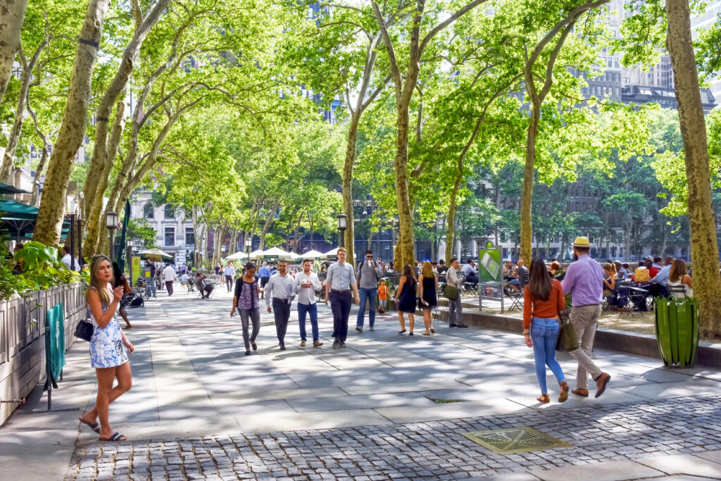 Photograph of a tree-lined park walkway filled with people walking toward and away from the camera, and people  sitting on benches to the right
