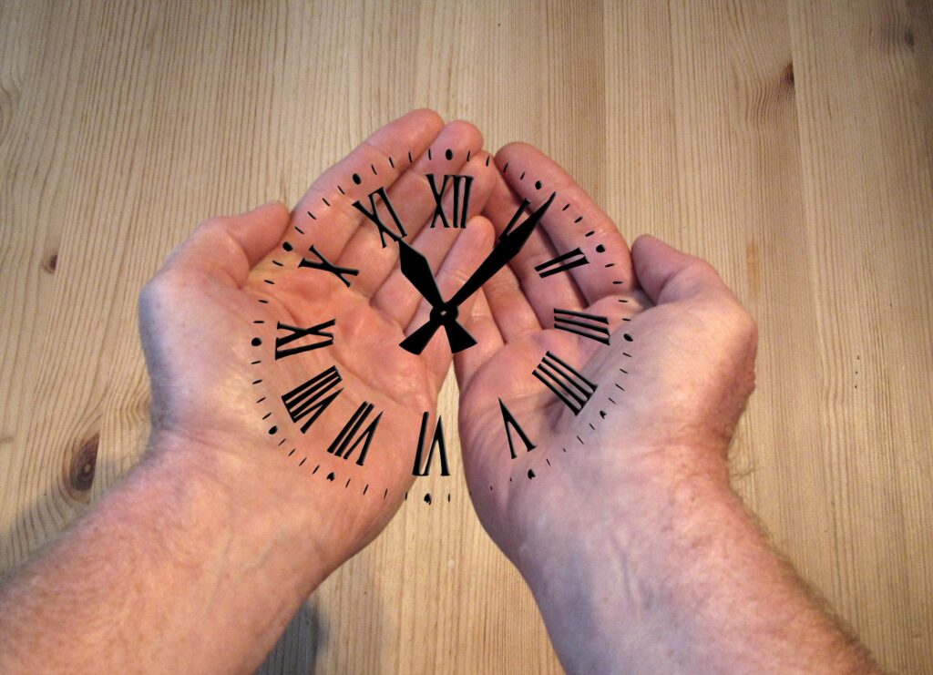 Photograph of a pale wood surface above which is a pair of cupped hands (male) with a clock face with roman numerals superimposed over the hands.