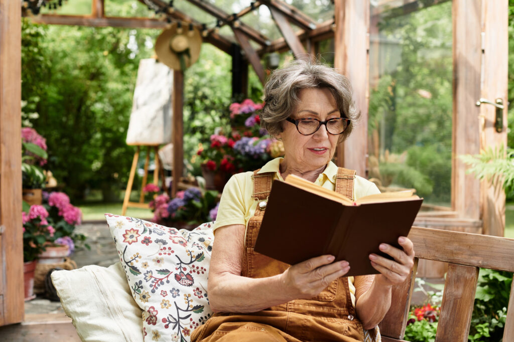 Photograph of a woman reading a book in a glasshouse garden filled with flowers with a treed landscape in the background