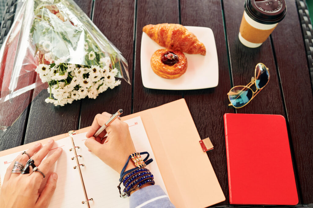 Photo from above looking down on a table with flowers, pastries, a cup of coffee, sunglasses, and a pair of women's hands with rings & bracelet, writing on a paper journal