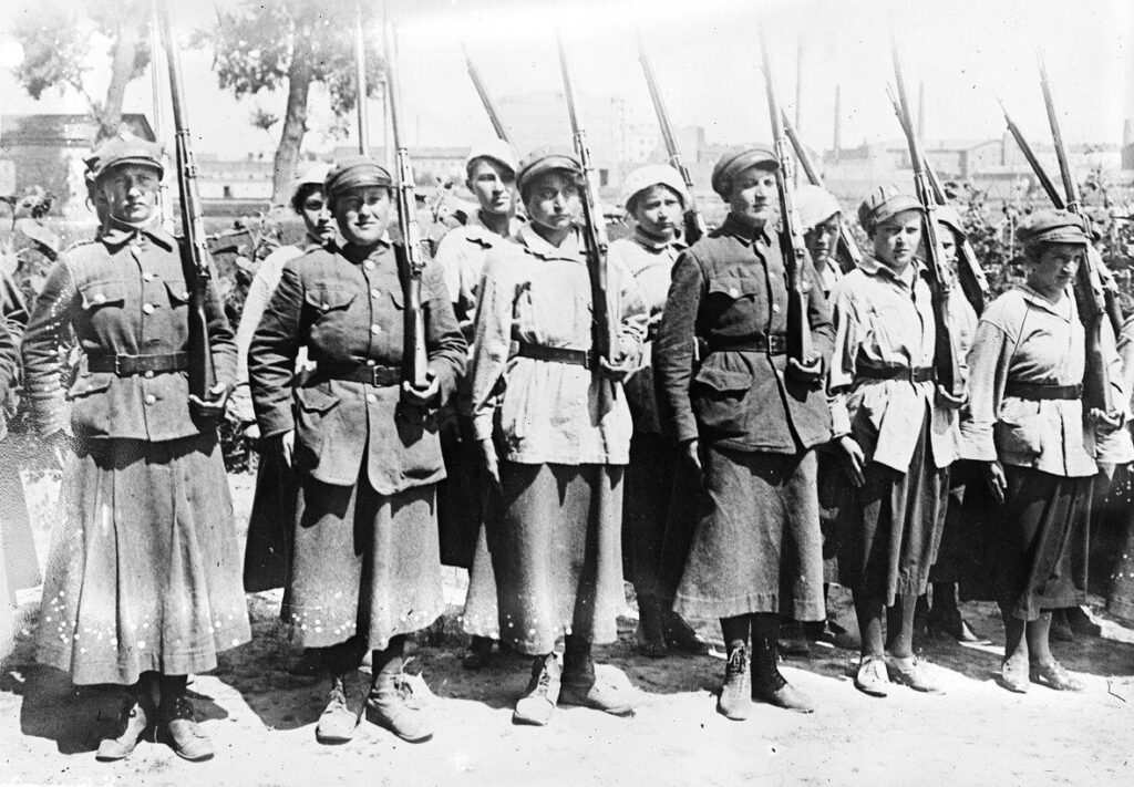 Black and white photograph of the women of 2nd Women’s Volunteer Legion. The women are in uniforms that are military caps, jackets, skirts, and boots and she woman shoulders a rifle with bayonet