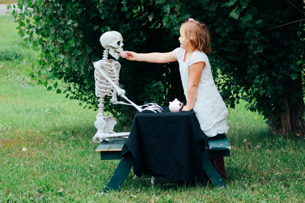 Photograph outdoors with a little girl at a child-sized table that holds a teapot, feeding her friend across the table who is a skeleton.