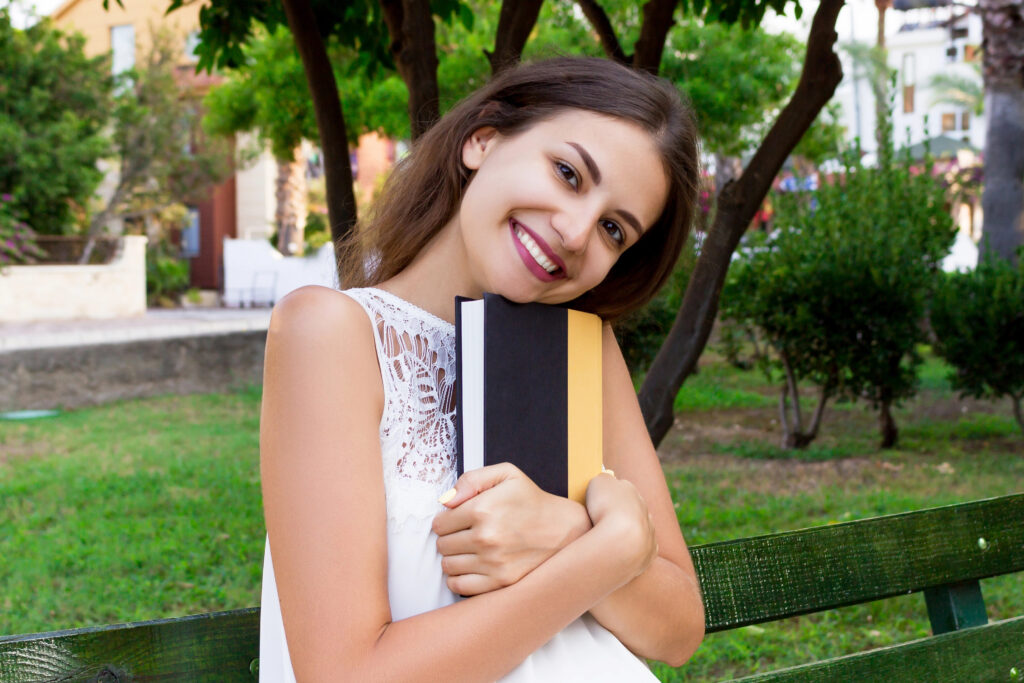 Photograph of a young woman standing outside infront of a green lawn dotted with trees and bushes, hugging a book, her head tilted. City buildings are in the background. 