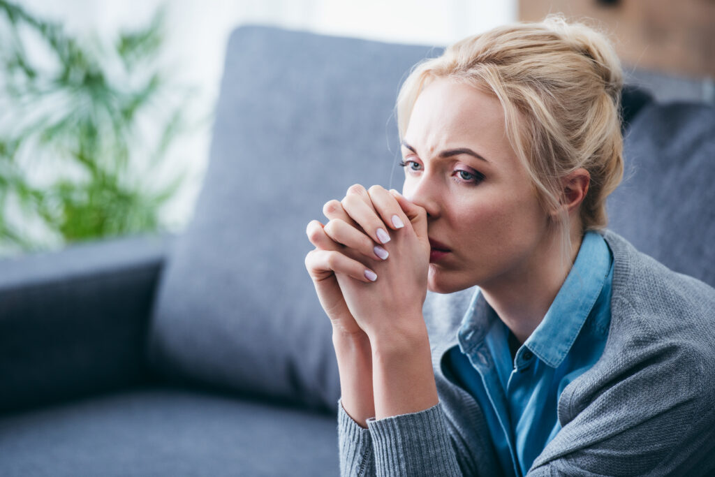 Photograph of a woman with a messy blonde bun, sitting on a blue-gray sofa, her hands folded together, her thumbs touching her nose and wearing a light gray sweater over a blue button down shirt.