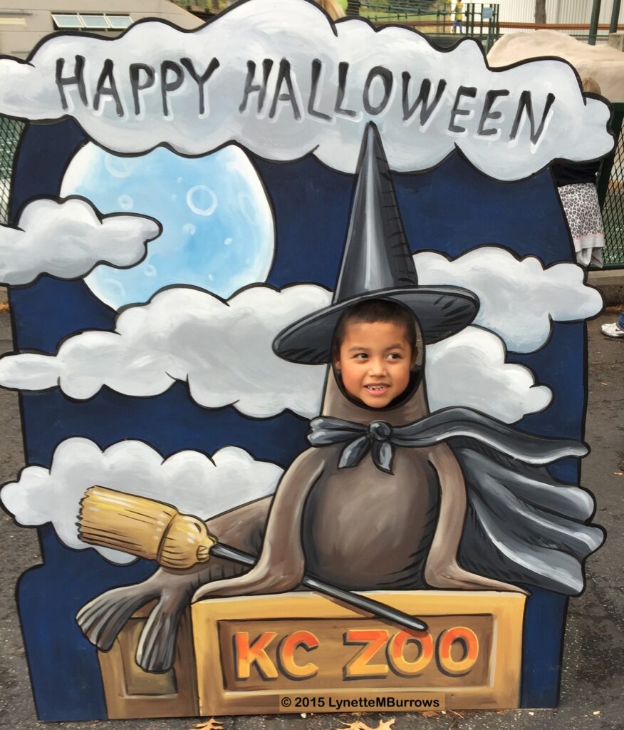 Photograph of a dark blue wooden sign decorated with Happy Halloween written across a cloud, more clouds and a moon, and a seal in a witches costume, the seal's head is a cut out and a small child's smiling face appears there.