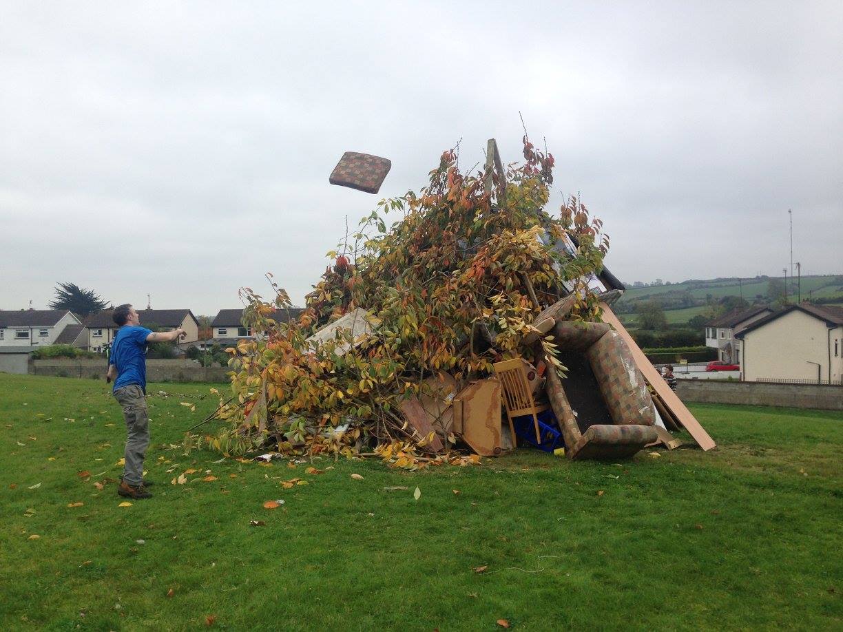 Photograph of a young man tossing a piece of wood or other combustable on top of a huge bonfire being built at Samhain in Ireland