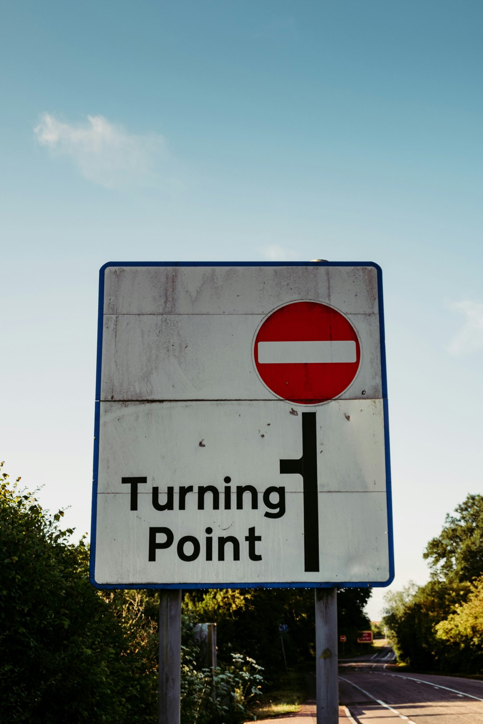 Photograph of a European road sign with a line indicating a road with a left hand turn, shortly after the turn is a red circle with a white horizontal bar across the center indicating dead end. The text on the sign reads "Turning point."